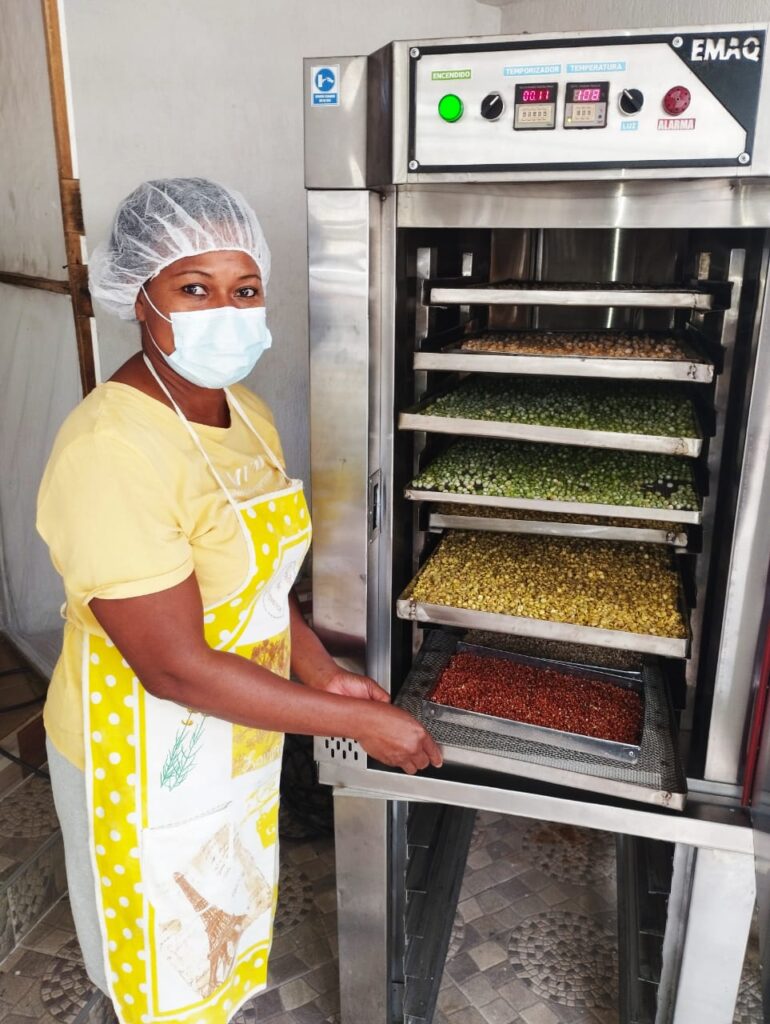 Mujer sonriente mientras seca legumbres en un horno deshidratador. Lleva puestos una cofia y un tapabocas.