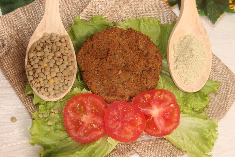 Al centro, carne de hamburguesa vegetal. A su izquierda, una cuchara de madera con lentejas, mienras a su derecha una chuchara de madera con harina de lentejas. alrededor decoraciòn con tomates tajados y hojas de lechuga.