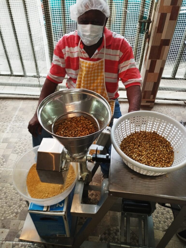 Hombre de tercera edad sonriendo mientras muele garbanzos. Lleva tapabocas y una cofia.