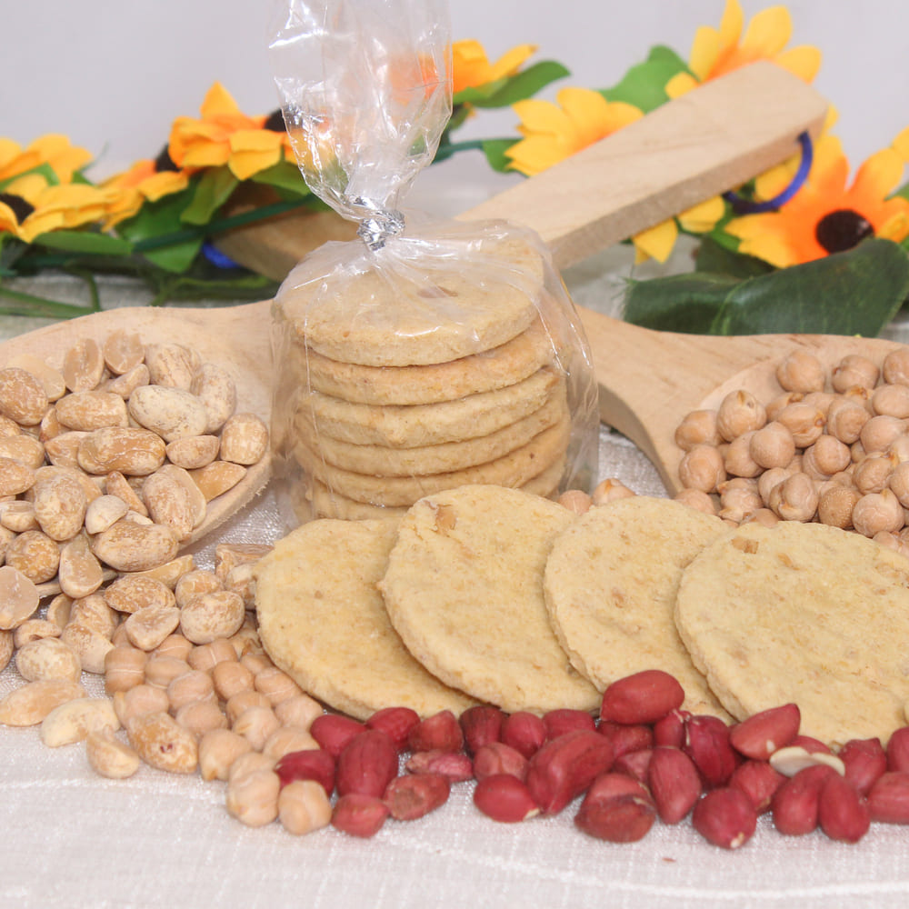 Al centro, paquete de 6 galletas de garbanzos y maní. Alrededor, decoración compuesta por galletas, garbanzos enteros en cucharas de madera y maní con cáscara. Al fondo, decoración de flores.