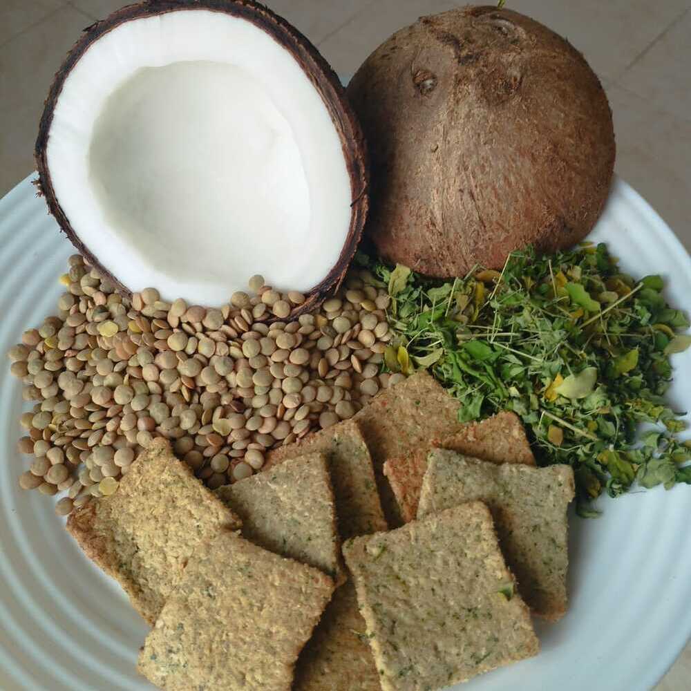 Biscotti quadrati di lenticchie, cocco e moringa. ai lati, lenticchie intere e foglioline di moringa verdi. Sullo sfondo, le due metá di un cocco.