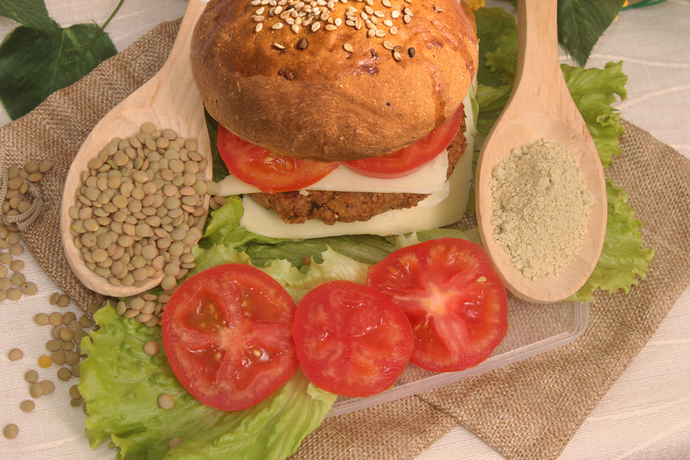 Hamburguesa con pan artesanal, tomate, queso, carne vegetal a base de lentejas y lechuga. Alrededor, dos cucharas de madera: la de la izquierda contiene lentejas enteras, la de la derecha contiene harina de lentejas precocidas.